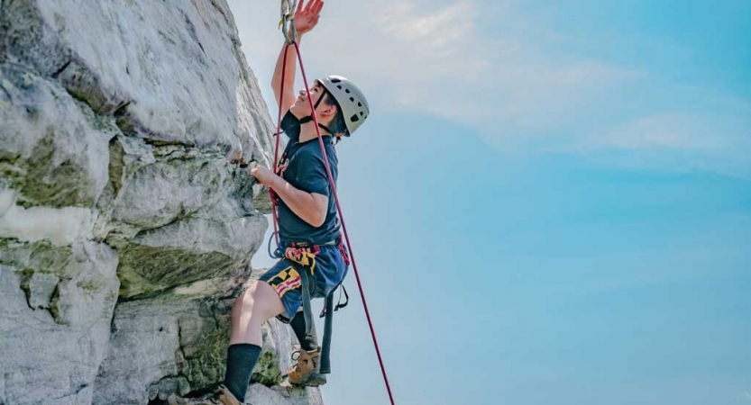 A young person wearing safety gear is secured by ropes as they reach higher on the wall they are climbing. The sky behind them is blue. 